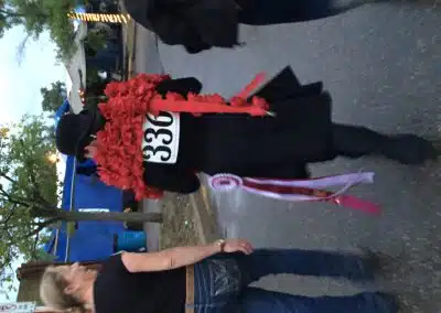 A person wearing a black coat, hat, and number 336, draped in red flowers and ribbons, walks outdoors with two others on a paved path near blue tents.
