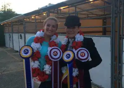 Two people stand side by side wearing flower garlands and holding award ribbons, with horse stalls in the background.