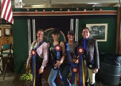 Four women stand indoors in front of a green wall, each holding award ribbons and smiling at the camera; an American flag hangs on the left.