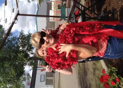 Woman wearing sunglasses and a large red flower lei smiles outdoors while another person behind her playfully makes bunny ears with their fingers.