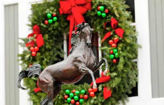 A bronze horse statue stands in front of a large holiday wreath decorated with red bows and green and red ornaments.